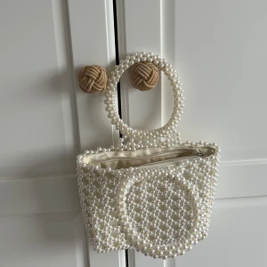 Vit pärlbeströdd handväska - Elegant handväska täckt med vita pärlor, perfekt för att ge en lyxig touch till din outfit. Väskan har ett runt handtag också dekorerat med pärlor och en dragkedja för säker förvaring. Perfekt för speciella tillfällen.