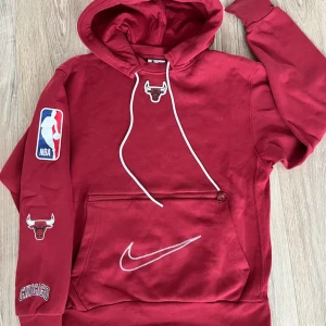 Röd hoodie från Nike med Chicago Bulls tema - Säljer en röd hoodie från Nike med Chicago Bulls logga på bröstet och ärmen. Hoodien har en stor ficka framtill med dragkedja och vita snören i huvan. Perfekt för en avslappnad stil. Ordinarie pris är 800kr