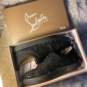 Svarta sneakers med nitar från Christian Louboutin - Säljer ett par svarta sneakers från Christian Louboutin med nitar på tån och sidorna. Skorna har en ikonisk röd sula och är tillverkade i mocka. Perfekta för att ge en edgy touch till din stil. Size 44