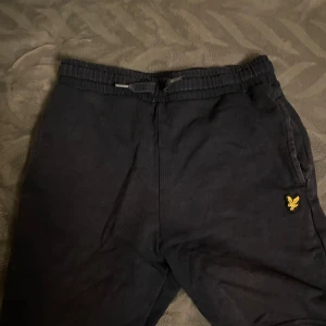 Blåa shorts från Lyle & Scott  - Säljer ett par blåa mjukisbyxor från Lyle & Scott Junior. Byxorna har en elastisk midja med snörning och en broderad logotyp på sidan. Perfekta för en bekväm och avslappnad stil.