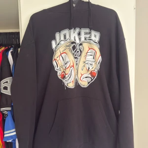 Joker brand  hoodie med clownmotiv - Säljer en svart hoodie med ett stort tryck av två clownansikten och texten 'Joker' på framsidan. Hoodien har en klassisk passform med långa ärmar och en rymlig ficka framtill. Perfekt för en avslappnad stil.