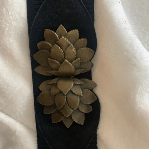 Svart bälte med dekorativa detaljer - Snyggt svart bälte med elastiskt material och dekorativa blommor i guld. Perfekt för att ge en extra touch till din outfit. Bältet har en bred design och är lätt att matcha med olika stilar.