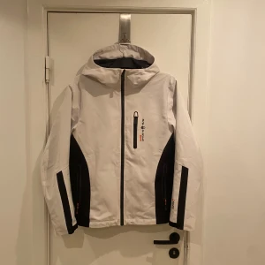 Vit och svart jacka från Sail Racing - Snygg vit och svart jacka från Sail Racing med dragkedja och huva. Jackan har flera praktiska fickor och en cool orange detalj på insidan. Passar Small bra! Skönt material och funkat perfekt som en lite tunnare jacka
