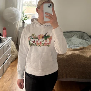 Vit hoodie från Levi's med blommigt tryck - Säljer en vit hoodie från Levi's med ett färgglatt blommigt tryck på bröstet. Hoodien har en klassisk passform med långa ärmar och en praktisk magficka. Perfekt för en avslappnad stil. Vid första tvätten åkte snörningen ur men den kan skickas med.