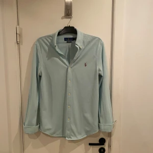 Ljusblå skjorta från Ralph Lauren - Snygg ljusblå skjorta från Ralph Lauren med klassisk krage och broderad logotyp på bröstet. Skjortan har långa ärmar och knappar framtill. Perfekt för en stilren look. Passar s perfekt. Riktigt skönt material