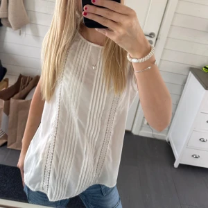 Vit blus med spetsdetaljer🤍 - Säljer en stilren vit blus med spetsdetaljer och veck framtill. Blusen är ärmlös och har en luftig passform, perfekt för varmare dagar. Passar både till jeans och kjol för en avslappnad look.