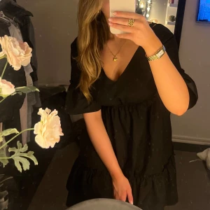 Svart klänning med öppen rygg - Snygg svart klänning med v-ringning och volangdetaljer. Den har en öppen rygg med snörning och korta ärmar. Perfekt för en kväll ute!   Helt oanvänd. (Sömmarna vid v-ringninhen är lite dåliga, var redan de när den köptes) 