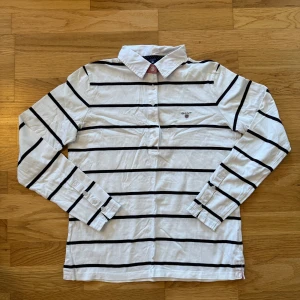 Vit och svart randig skjorta från Gant - Säljer en stilren vit piké från Gant med svarta ränder. Skjortan har långa ärmar och knappar framtill samt en klassisk krage. Perfekt för en avslappnad men ändå uppklädd look till sommaren. Säljer pga för liten för mig.