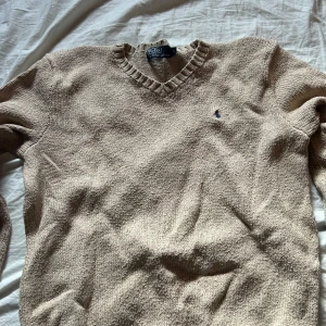 Beige stickad tröja från Polo Ralph Lauren - Säljer en klassisk beige stickad tröja från Polo Ralph Lauren med v-ringning och det ikoniska logotypbroderiet på bröstet. Perfekt för kyligare dagar med sin mjuka och bekväma passform. Passar både till jeans och chinos.