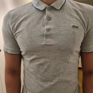 Grå pikétröja från Lacoste - Grå pikétröja från Lacoste med klassisk krage och tre knappar. Tröjan har korta ärmar och det ikoniska krokodilmärket broderat på bröstet.