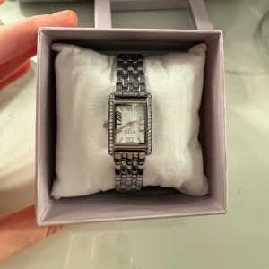 Silverfärgad klocka från Regal - Elegant silverfärgad klocka från Regal med rektangulär urtavla och romerska siffror. Klockan har en glittrig ram runt urtavlan och en länkarmband i metall. Perfekt för att ge en stilren touch till din outfit.