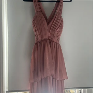 Rosa långklänning med v-ringning - Elegant rosa långklänning med v-ringning och veckad överdel. Perfekt för festliga tillfällen. Klänningen har en flytande kjol som ger en vacker silhuett.