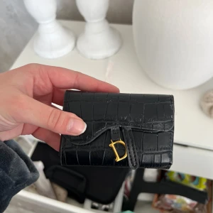 Svart plånbok med krokodilmönster - Snygg svart plånbok i skinn med krokodilmönster och gulddetaljer. Den har en praktisk design med flera fack för kort och en säker stängning med en knapp. Perfekt för att hålla ordning på dina kort och kontanter.