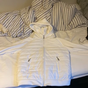 Vit quiltad väst med huva - Snygg vit quiltad väst med huva och dragkedja framtill. Västen har två praktiska fickor med dragkedjor. Perfekt för lager-på-lager under kyligare dagar.