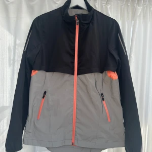Svart och grå vindjacka från Cubus - Säljer en stilren svart och grå vindjacka från Cubus i storlek M. Jackan har en dragkedja i orange och matchande detaljer på fickorna. Tillverkad i 100% polyester med långärmad design och reflekterande element på fram och baksida.