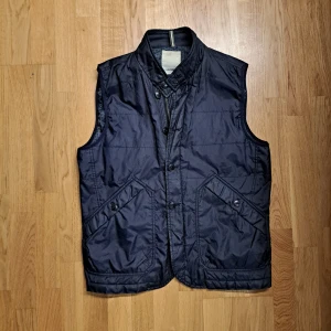 Marinblå väst från J.Lindeberg - Snygg marinblå väst från J.Lindeberg med knappar och dragkedja. Perfekt för lager-på-lager stil. Västen har en stilren design med två fickor framtill. Skriv vid fler frågor