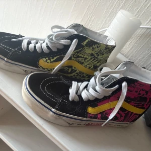Svarta sneakers med graffiti-mönster - Coola svarta sneakers från Vans med graffiti-mönster i rosa och gul. Skorna har en hög modell med vit snörning och en gul sidostrimma. Perfekta för att ge din outfit en edgy touch.