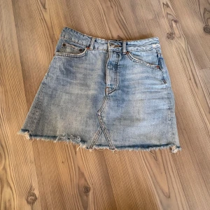 Ljusblå jeanskjol med rå kant - Trendig ljusblå jeanskjol med rå, fransig kant och klassiska fem fickor. Kjolen har knappgylf framtill och en lätt A-linjeformad siluett. Perfekt för dig som gillar en avslappnad och cool stil.