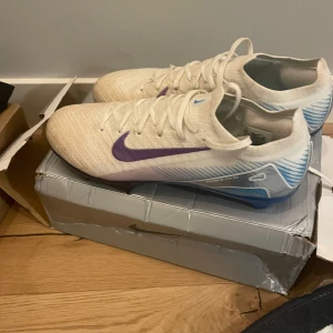 Fotbollsskor - Säljer ett par vita Nike Air fotbollsskor med lila swoosh och blå detaljer. Skorna har snörning och en rund tå. Yttermaterialet är syntet och sulan har blått mönster. Perfekta för dig som vill sticka ut på planen!