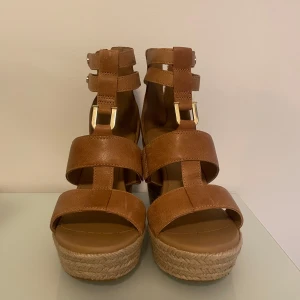 Bruna kilklackade sandaler från UGG  - Snygga bruna sandaler från UGG med hög kilklack i flätad jute. Skorna har breda remmar, guldfärgade detaljer och dragkedja baktill. Perfekta för sommaren och ger en bohemisk touch till din outfit. Modell Kolfax Almond. Nypris 1049 kr. Aldrig använda! 