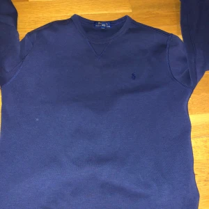 Blå tröja från Polo Ralph Lauren - Mörkblå tröja från Polo Ralph Lauren med diskret broderad logga på bröstet. Klassisk v-ringad modell med långa ärmar och mjukt material. Perfekt för en stilren och enkel look. Storlek 14-16 år