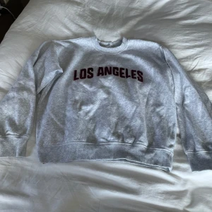 Grå sweatshirt med Los Angeles-tryck - Säljer en grå sweatshirt med texten 'Los Angeles' i vinrött på bröstet. Tröjan har långa ärmar och ribbade muddar vid ärmslut och nederkant. Perfekt för en avslappnad stil!