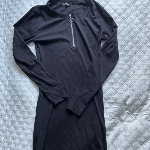 Svart ribbad klänning med dragkedja - Snygg svart ribbad klänning med lång ärm och en cool dragkedja framtill. Perfekt för en stilren look.