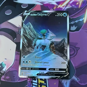 Pokémonkort Ice Rider Calyrex V - Säljer ett Pokémonkort: Ice Rider Calyrex V med 210 HP. Kortet har en isblå och silver design med motiv av Calyrex på en ishäst framför snötäckta berg. Kortet har attacker som Pierce och Glacial Lance. Perfekt för samlare eller Pokémonspelare.