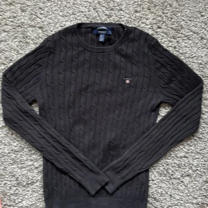 Svart kabelstickad tröja från GANT - Säljer en svart kabelstickad tröja från GANT i stretchig bomull. Tröjan har rund halsringning, långa ärmar och diskret GANT-logga broderad på bröstet. Perfekt för en stilren och klassisk look.