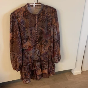 Mönstrad volangklänning i lila och bruna gula toner - Säljer en unik klänning med volanger och knappar framtill. Klänningen har lång ärm och ett färgstarkt paisleymönster i lila, brunt och orange. Perfekt för dig som gillar bohemisk stil och vill sticka ut med en flowy känsla. Ha linne under. Inbyggd svart kjol