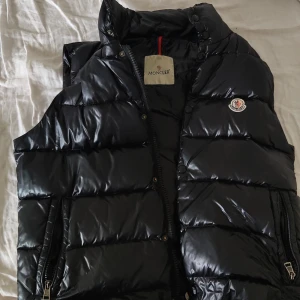Svart dunväst från Moncler - Säljer en svart dunväst från Moncler med hög krage och dragkedja framtill. Västen har två fickor med dragkedja och Moncler-logga på bröstet. Perfekt för lager-på-lager under kyliga dagar.