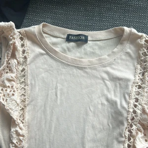 Beige topp med spetsdetaljer från Fashion - Säljer en beige topp från Fashion med dekorativa spetsvolanger längs axlarna och sidorna framtill. Toppen har rund halsringning och är kortärmad. Perfekt för dig som gillar romantiska detaljer!