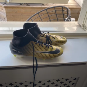 Nike Mercurial fotbollsskor i gult och svart - Säljer ett par Nike Mercurial fotbollsskor i gult med svarta detaljer och vita ränder. Skorna har hög krage och snörning för extra stöd. Sulan har fasta dobbar i gult och vitt, perfekt för spel på gräsplan. Kolla in min profil för mer.