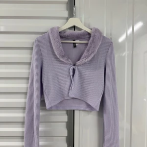 Ljuslila croppad kofta från Divided - Superfin ljuslila croppad kofta från Divided med mjuk fuskpäls runt kragen och ribbad struktur. Långa ärmar och knyt framtill ger en trendig look. Perfekt att styla med jeans eller kjol för en cool vibe.