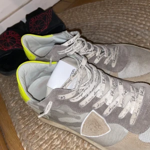 Philippe Model sneakers med camomönster - Snygga sneakers från Philippe Model i beige och grå toner med camouflagemönster och detaljer i mocka. Skorna har snörning, vit sula och en gul detalj vid hälen. Perfekta för dig som gillar streetstyle och unika detaljer.