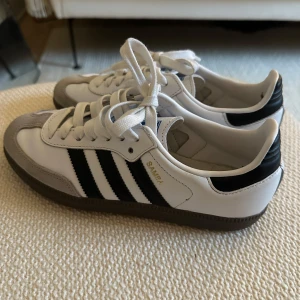 Adidas Samba sneakers - Klassiska Adidas Samba sneakers med vit ovandel, grå mockadetaljer och svarta ränder på sidorna. Perfekta för en sportig och stilren look. Storlek 36 2/3 - passar mig med 37. Använda 2-3 gånger och inköpta förra året. 🖤