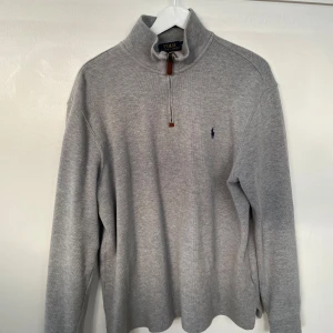 Grå halvzip tröja från Polo Ralph Lauren - Säljer en klassisk grå långärmad tröja från Polo Ralph Lauren med halv dragkedja och hög krage. Tröjan har en liten broderad logga på bröstet och är tillverkad i mjukt material. Perfekt för lager-på-lager-stil.