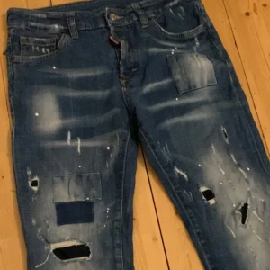 Dsquared2 Cool Guy Jeans med slitningar och lappar - Säljer ett par blå Dsquared2 Cool Guy Jeans med tydliga slitningar, hål och lappar på benen. Jeansen har klassisk femficksdesign och knappgylf. Snyggt tvättade detaljer och patchar ger en unik look.