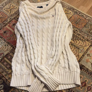 Vit kabelstickad tröja från GANT - Säljer en klassisk vit kabelstickad tröja från GANT med rund halsringning och diskret logga broderad på bröstet. Tröjan har långa ärmar och ribbade muddar vid ärmslut och nederkant. Perfekt för en stilren och tidlös look.