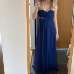 Blå långklänning med pärldetaljer - Elegant marinblå långklänning med vackra pärldetaljer och veckad överdel. Klänningen är axelbandslös och har en markerad midja med en fin detalj vid bröstet.. Perfekt för bal eller festliga tillfällen. Mycket bra skick!! 