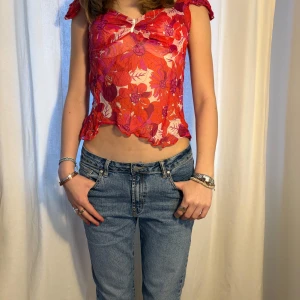Blommig mesh topp i rött och rosa - Säljer en blommig mesh topp i rött och rosa med vita detaljer. Toppen har korta ärmar, v-ringning och är croppad med vågig nederkant. Perfekt för dig som gillar färg och mönster!
