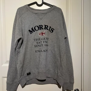 Grå sweatshirt från Morris - Grå sweatshirt från Morris med broderad text och brittisk flagga på bröstet. Tröjan har rund halsringning, långa ärmar och ribbade muddar vid ärmslut och nederkant. Perfekt för en avslappnad och stilren look.