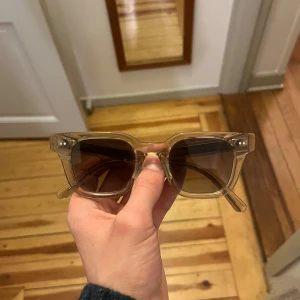 CHIMI Glasögon - Snygga solglasögon från CHIMI med transparent beige båge och glittrig gulddetalj på ovansidan. Fyrkantig form och mörka glas. Perfekt accessoar för att lyfta din stil.