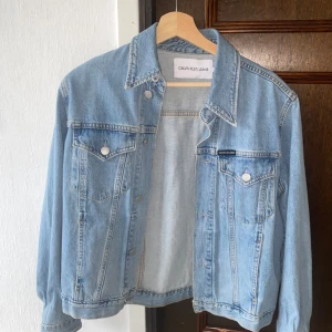 Ljusblå jeansjacka från Calvin Klein Jeans - Säljer en klassisk ljusblå jeansjacka från Calvin Klein Jeans. Jackan har två bröstfickor med lock och knappar, samt en rak passform och krage. Perfekt för dig som vill bära något enkelt och fint över din outfit.