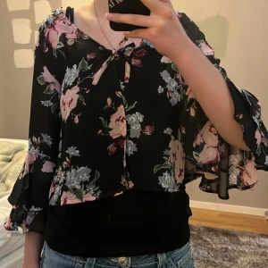 Super söt blommig blus med volangärmar - Säljer en svart blus med blommönster. Blusen har vida volangärmar och knytning vid halsen. Tunt, lätt material som ger ett luftigt intryck. Passar perfekt med ett linne under! Super fin till fest men även sommarkvällar, super söt och stilren 🌺🌸🌻🌼🖤
