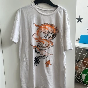 Vit t-shirt med draktryck - Vit t-shirt med stort, detaljerat drakmotiv i orange och svart på framsidan. T-shirten har rund hals och korta ärmar. Perfekt för dig som gillar streetstyle och vill sticka ut med ett coolt print. Oversized 