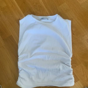 Vit ärmlös topp från Zara - Säljer en stilren vit ärmlös topp från Zara. Toppen har en enkel design med rynkade detaljer på sidorna som ger en snygg touch. Köparen står för frakt 