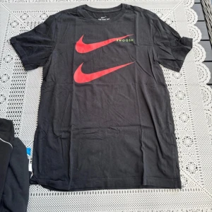 Svart t-shirt från Nike med röd logga - Säljer en svart t-shirt från Nike med två röda swoosh-loggor på bröstet. T-shirten är kortärmad och har en klassisk rund halsringning. Perfekt för en sportig look eller avslappnad stil.