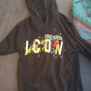 Svart hoodie från Dsquared2 med färgstänk - Säljer en svart hoodie från Dsquared2 med stor vit 'ICON'-text och färgglada färgstänk i gult, rött, blått och grönt på framsidan. Tröjan har huva och lång ärm, perfekt för en cool och avslappnad stil. 