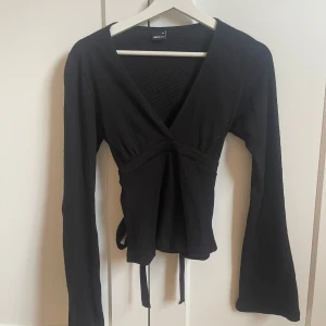 Svart topp med knyt från Gina Tricot - Säljer en svart topp från Gina Tricot i storlek XS. Toppen har långa ärmar, v-ringning och knytband i midjan för en snygg passform. Perfekt att styla med jeans eller kjol💗